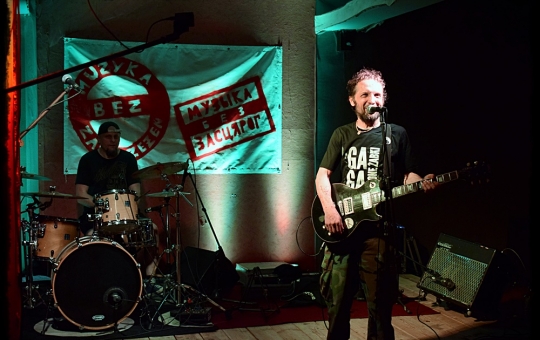 Na zdjęciu znajdują się muzycy zespołu Ga-ga-Zielone żabki, z przodu przy mikrofonie stoi wokalista z gitarą, za nim gra perkusista, w tle widać baner z napisem Muzyka bez zastrzeżeń.