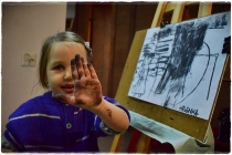 Na zdjeciu widnieje dziewczynka, która pokazuje brudną od wękla dłoń. Obok stoi sztaluga z namalowanym węglem rysunkiem.