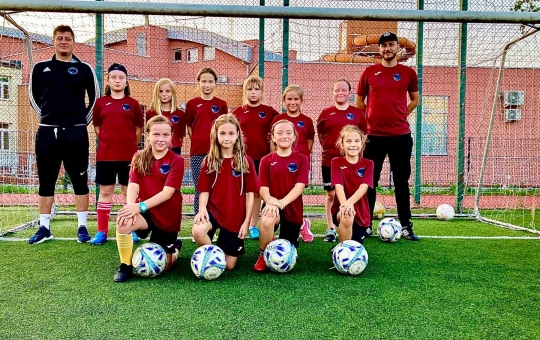 Drużyna dziewcząt i trenerzy stoją na tle bramki do piłki nożnej. Wszyscy ubrani w klubowe stroje Pasji