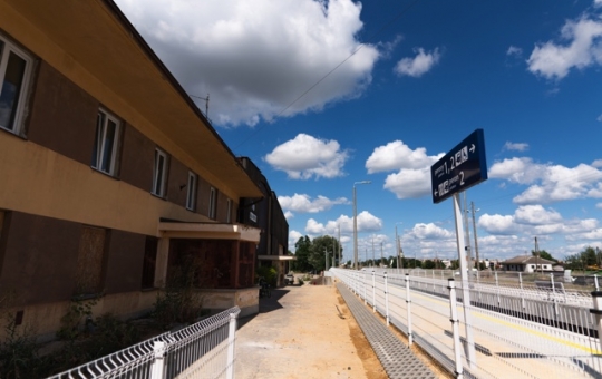 zdjęcie: z lewej strony front budynku dworaca PKP w Hajnówce, po prawej stronie tory kolejowe, nad nimi unosi się błękitne niebo z obłokami