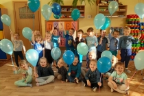 Zdjęcie przedstawia grupę dzieci pozującą do zdjęcia. Część z nich stoi, a część siedzi na podłodze w przedszkolnej sali. Wszystkie trzymają w dłoniach niebieskie balony.