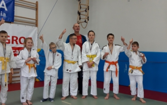 Zdjęcie przedstawia siedmioosobową grupę judoków z Hajnówki wraz z trenerem. Zawodnicy ubrani są w białe kimona przepasane żółtymi pasami. Za szyi zawieszone maja medale.