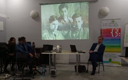 Po prawej stronie zdjęcia siedzi Wojciech Konończuk, z lewej zezpół Zbigniewa Budzyńskiego. W centrum widnieje duże czarno-białe zdjęcie wyświetlane na rzutniku.