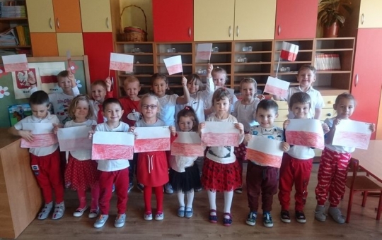 Zdjęcie przedstawia przedszkolaki z grupy Sówki. Dzieci stoją w dwóch rzędach w przedszkolnej sali, w rękach trzymają rysunki flagi Polski.