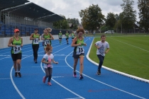Dzieci, młodzież i dorośli biegną w biegu dla Kubusia. Wielu z nich ubranych jest w zielone koszulki klubu Hajnówka Biega. W tle widać trybuny i zieloną murawę
