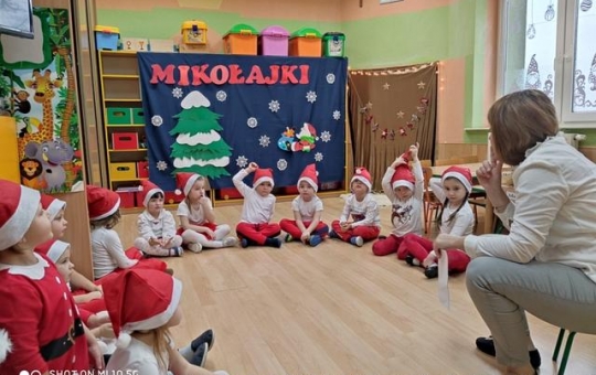 Przedszkolaki siedzą na podłodze, przed nimi na krześle siedzi wychowawczyni. Dzieci ubrane sa w ubrania w kolorach biało - czerwonych, na głowach maja czapki mikołaja.
