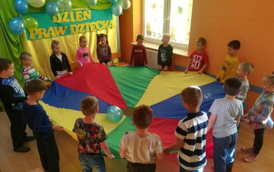 Grupa dzieci stojąc w kole trzyma chustę Klanzy bawiąc się balonem.