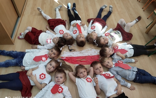 Grupa dzieci ubranych w stroje galowe leży na podłodze w kręgu stykając się głowami. W środku ułożona jest flaga Polski z białej i czerwonej bibuły.