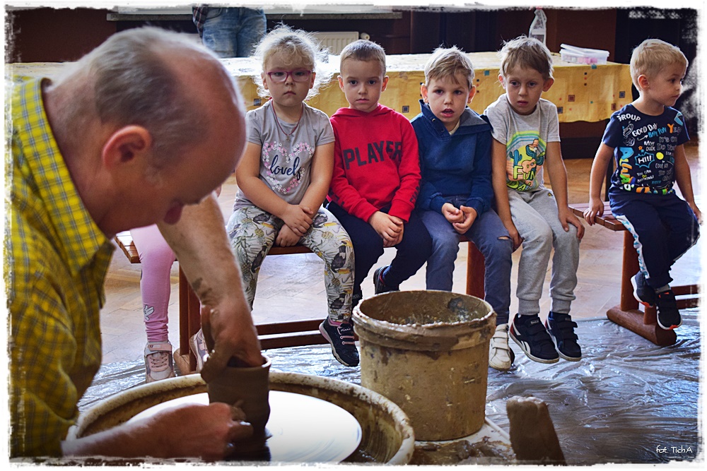 Na zdjęciu prowadzący pokazuje jak na kole powstają naczynia z gliny. W tle siedzi sześcioro dzieci, które przyglądają się pracy garncarza.
