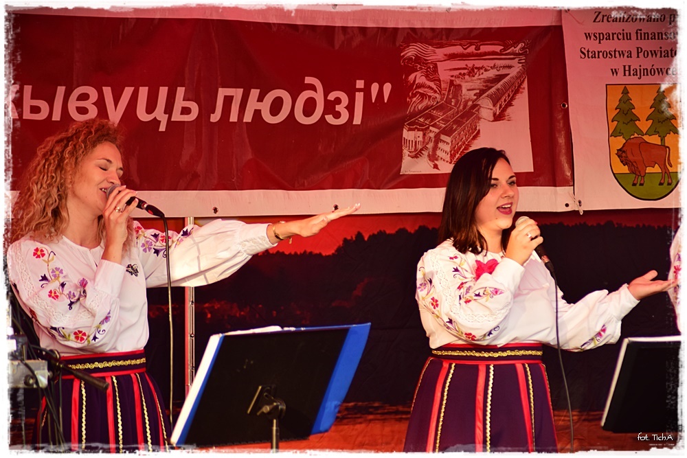 Na zdjęciu dwie śpiewające wokalistki ubrane w ludowe stroje. W tle fragment białoruskiego napisu i herb Startostwa Powiatowego w Hajnówce.