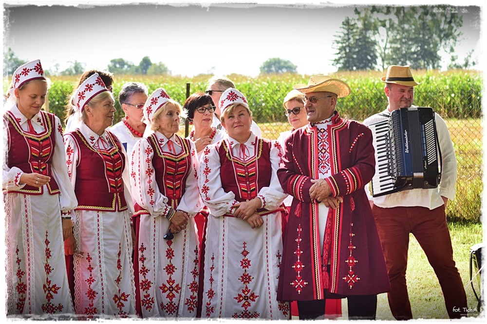 Na zdjęciu białoruski chór w tradycyjnych strojach. Z prawej strony stoją dwaj mężczyźni w kapeluszach. Jeden ma akordeon. W tle widać pole.