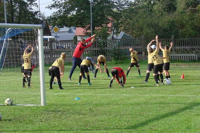 Młodzi piłkarze stoją na marawie boiska i rozgrzewaja się przed meczem. Cześć z nich ma ręcę uniesione dogóry, część wykonuje skłon.
