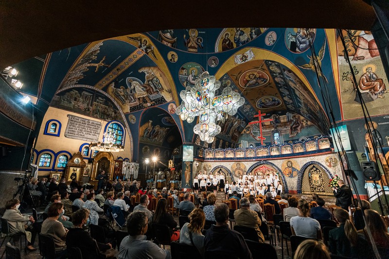 Zdjęcie przedstawia piękne wnętrze cerkwii, w której publiczność słucha chóru dziecięcego. Nad publicznością zawieszony jest duży szklany krzyż. Całe wnętrze cerkwii pokryte jest freskami.