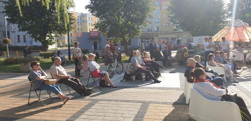 Zgromadzeni mieszkańcy Hajnówki, stoją lub siedzą na krzesłach, w tle drzewa i ul. 3 Maja