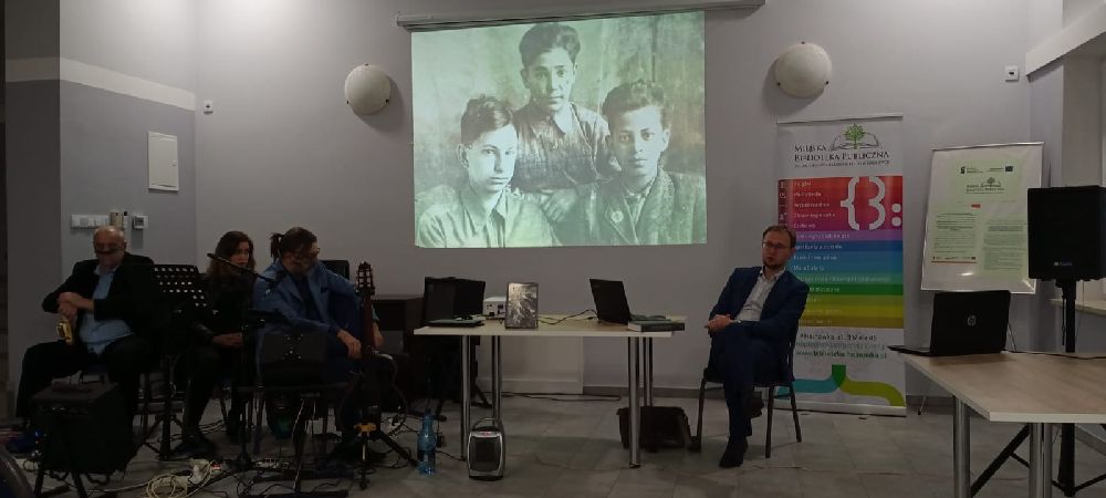 Po prawej stronie zdjęcia siedzi Wojciech Konończuk, z lewej zezpół Zbigniewa Budzyńskiego. W centrum widnieje duże czarno-białe zdjęcie wyświetlane na rzutniku.