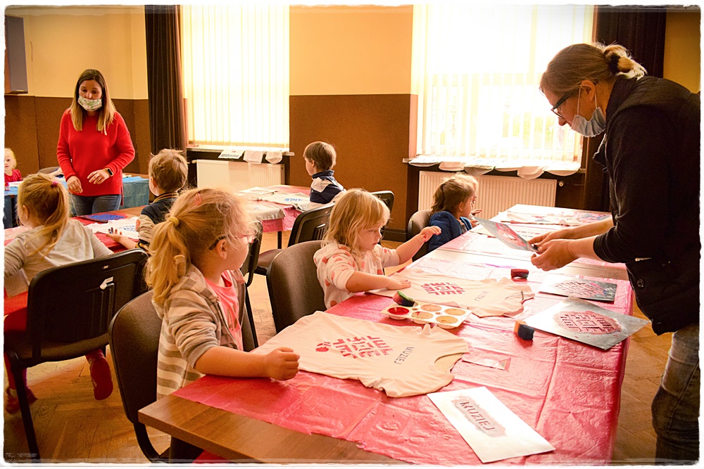 Zdjęcie przedsztaweiające dzieci uczestniczące w warsztatach odbijania szablonami wzory białoruskie na koszulkach. siedmioro dzieci siedzi przy stołach, dwie opiekunki stoją obok.