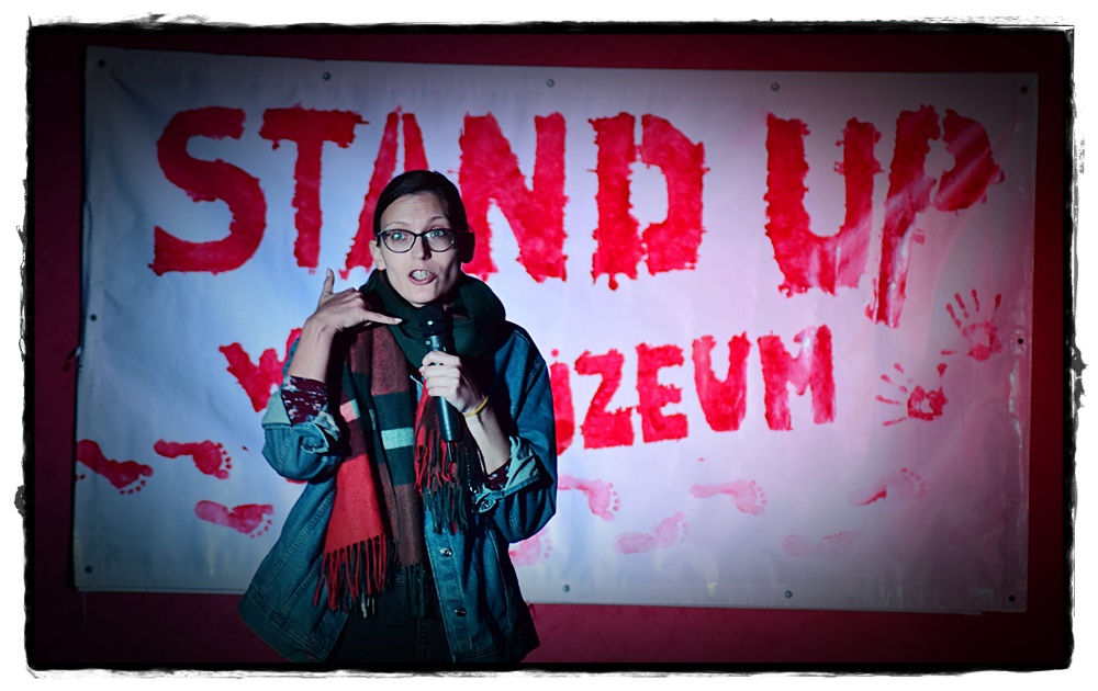 W centrum zdjecia stoi kobieta, trzyma w rekach mikrofon.W tle wisi duży biały baner z czerwonym napisem: Stand up muzeum.