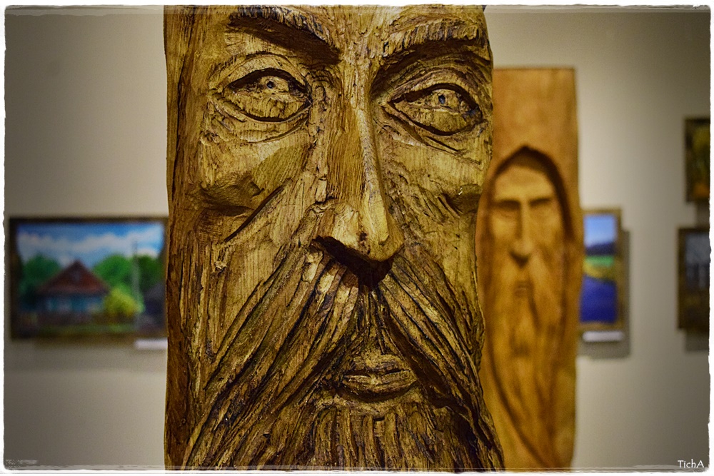 Na zdjęciu widnieje fragment rzeźby w drewnie - męska twarz. W tle widać inną rzeźbę przedstawiającą starca oraz zawieszone obrazy.