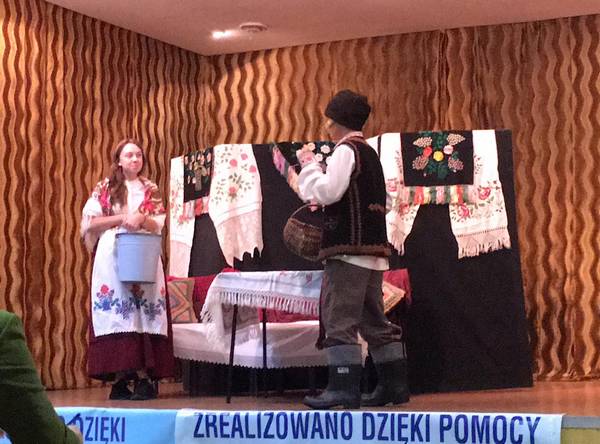 Na scenie BTSK stoi dziewczyna z wiadrem i chłopak z koszykiem obróceni twarzą do siebie, ubrani są w ludowe stroje. na scenie znajduje się również dekoracja wykonana z ręczników i makatek białoruskich.