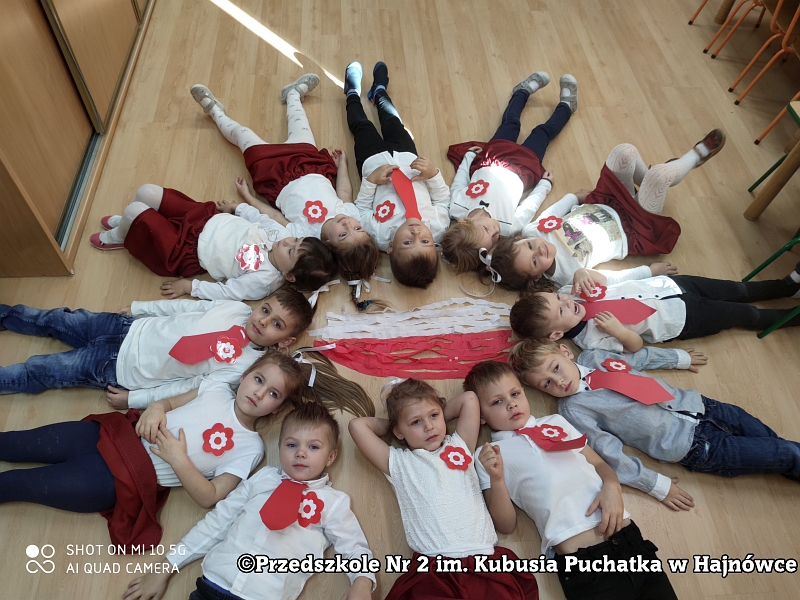 Grupa dzieci ubranych w stroje galowe leży na podłodze w kręgu stykając się głowami. W środku ułożona jest flaga Polski z białej i czerwonej bibuły.