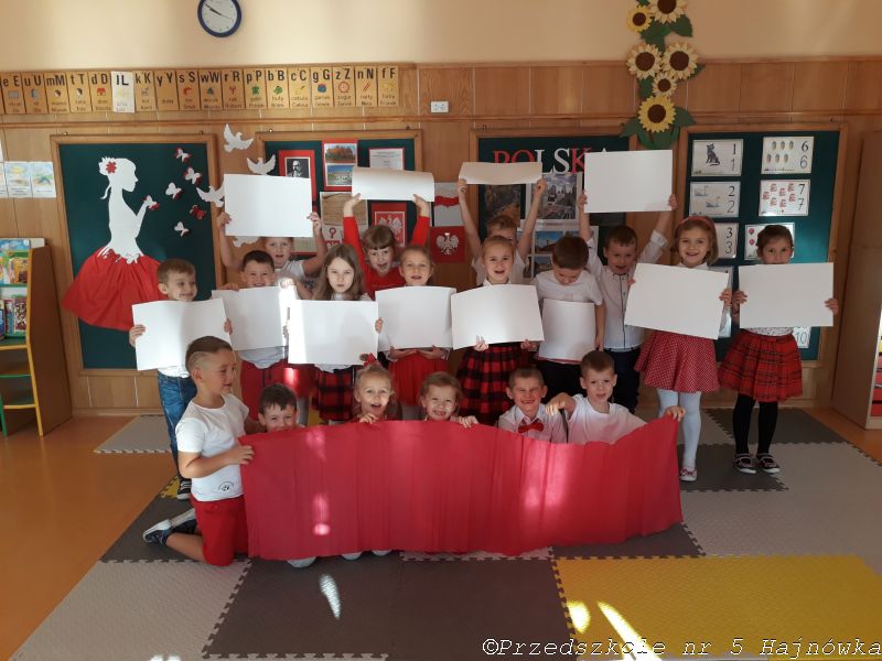 Grupa dzieci ubranych w biało-czerwone stroje trzyma kartki w tych samych kolorach,tworząc flagę Polski. Z tyłu wisi gazetka okazjonalna.