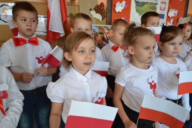Zdjęcie prezentuje ośmioro dzieci z grupy Sarenek podczas recytacji wiersza „Dzień dobry biały ptaku”. Przedszkolaki są ubrani na galowo, przy białych bluzkach mają przypięte biało-czerwone kotyliony a w rękach trzymają chorągiewki.