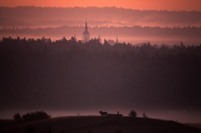 Zdjęcie przedstawia widok na wieżę kościoła za tle zachodzącego słońca. Na pierwszym planie pagórek, na nim koń z zaprzęgniętą furą i postacią.