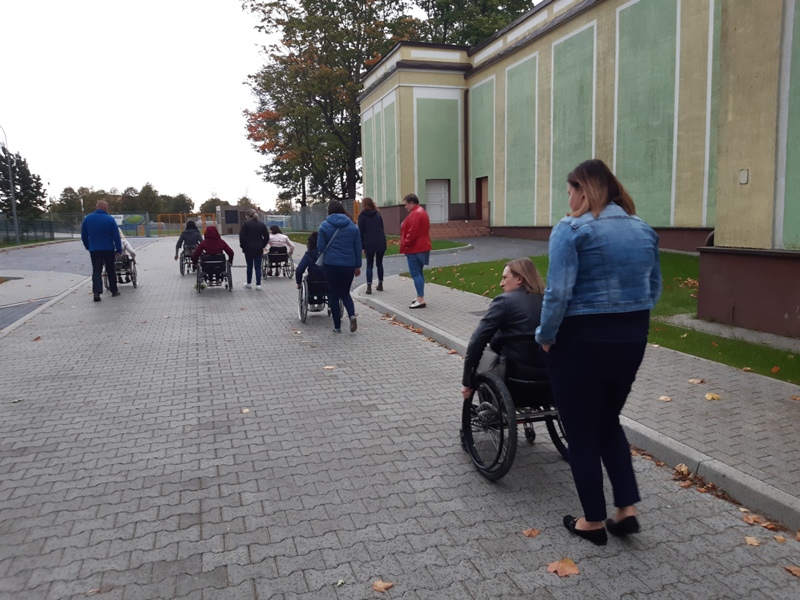 zdjęcie: podwórze; 6 osób na wózkach inwalidzkich, za nimi osoby pełniące role ich asystentów; poruszają się przed siebie po ścieżce z szarej kostki brukowej