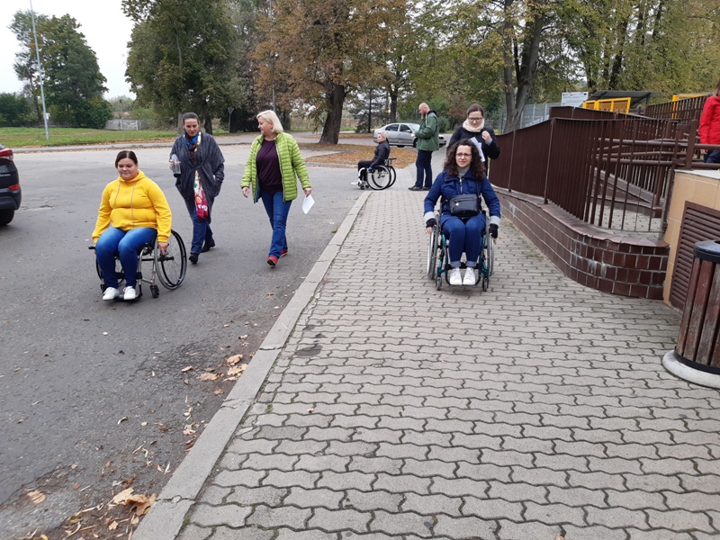 zdjęcie: podwórze; trzy osoby na wózkach (kobiety) i cztery idące z nimi (trzy kobiety i jeden mężczyzna); z prawej strony znajduje się podjazd dla osób z niepełnosprawnościami