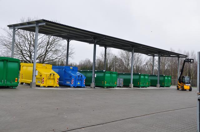 Zdjęcie przedstawia żółte, niebieskie i zielone pojemniki na odpady umieszczone pod zadaszona wiatą. Po prawej stronie stoi wózek widłowy.
